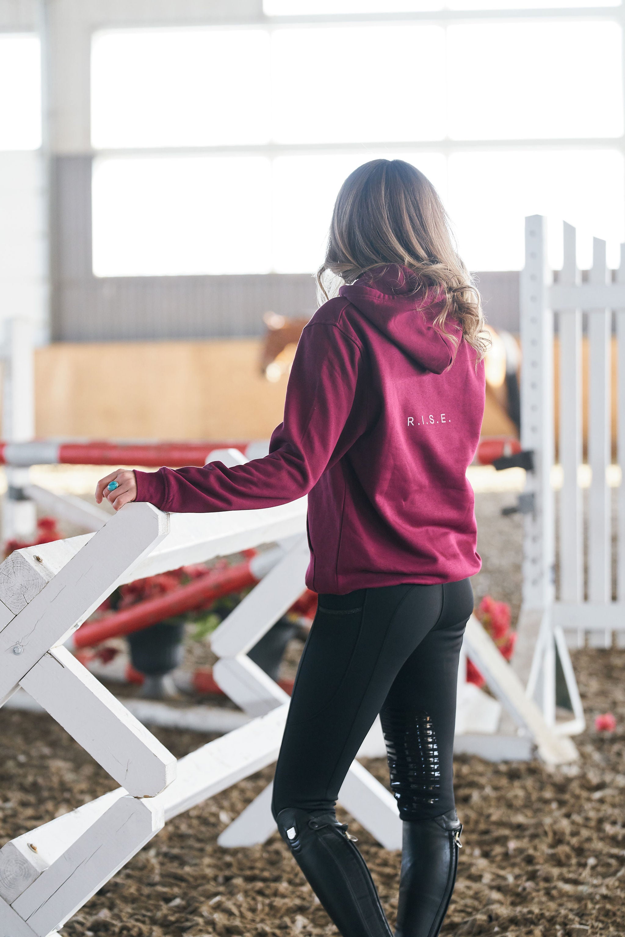 Person wearing a maroon hoodie with 'RISE branding in an equestrian setting.